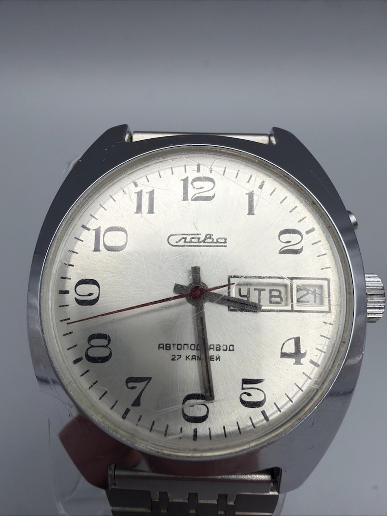 May include: Silver-toned wristwatch with a round face and a silver band. The watch face has black numerals, a day-date display, and the word "Слава" in Cyrillic script. The watch hands are red.