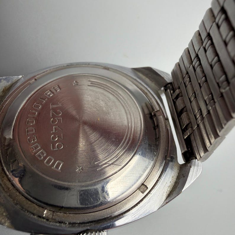 May include: Close-up of a vintage silver-toned wristwatch. The watch back features engraved text and the number 125459. The watch band is a woven metal mesh. The watch has a circular face with a textured design.