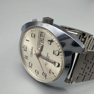 May include: A vintage silver-toned wristwatch with a cream-colored face. The watch features black numerals, a day-date display, and a red second hand. The watchband is a silver-toned, mesh-style design.