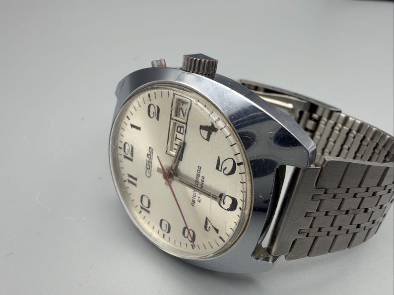 May include: A vintage silver-toned wristwatch with a cream-colored face. The watch features black numerals, a day-date display, and a red second hand. The watchband is a silver-toned, mesh-style design.