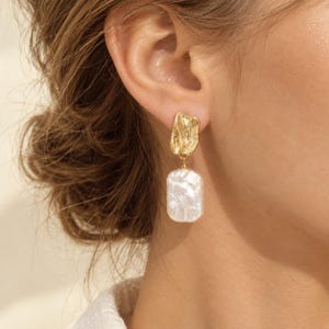 May include: A gold-toned earring featuring an irregular, textured metal stud at the top connected to a rectangular, white, baroque-style pearl drop. The metal component has a rippled, organic surface finish, while the pearl displays a lustrous, uneven texture.