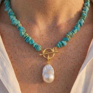 May include: A necklace featuring a strand of irregular, bright blue turquoise chips. The necklace includes a gold-toned toggle clasp at the centre, from which hangs a single, large, white, irregular-shaped baroque pearl pendant.