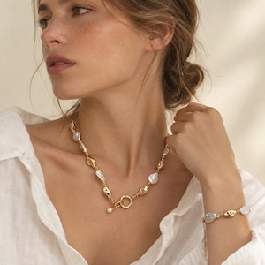 May include: A gold-toned chain necklace and matching bracelet set featuring alternating irregular white baroque pearls and smooth, organic-shaped gold-toned metal beads. The necklace includes a circular toggle clasp closure at the front.