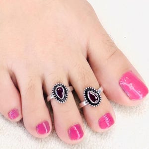Peut inclure: Deux bagues d'orteil en argent avec une pierre rouge foncé en forme de goutte sertie dans un cadre en métal perlé. Chaque bague est portée sur un orteil, présentant une finition métallique polie avec des détails complexes autour du sertissage.