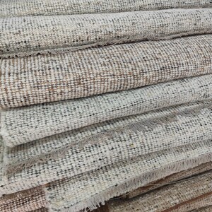 May include: A stack of multiple rectangular woven rugs featuring a textured, multi-colored pattern of cream, beige, gray, and brown fibers. Each rug is tightly rolled or folded, displaying visible frayed fringe edges along the horizontal borders.
