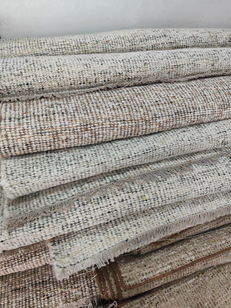 May include: A stack of multiple rectangular woven rugs featuring a textured, multi-colored pattern of cream, beige, gray, and brown fibers. Each rug is tightly rolled or folded, displaying visible frayed fringe edges along the horizontal borders.