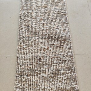 May include: A rectangular runner rug featuring a thick, hand-woven texture with a blend of neutral tones including beige, tan, cream, and white. The surface displays a chunky, uneven weave with scattered tufts of white yarn throughout the design.