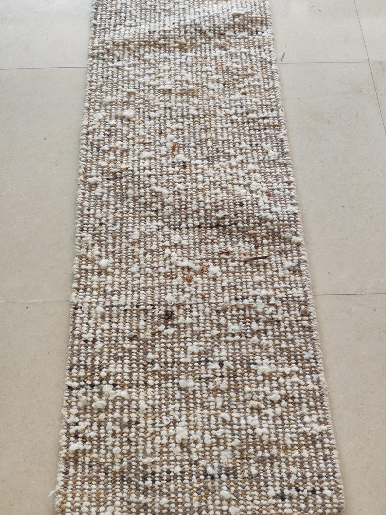 May include: A rectangular runner rug featuring a thick, hand-woven texture with a blend of neutral tones including beige, tan, cream, and white. The surface displays a chunky, uneven weave with scattered tufts of white yarn throughout the design.