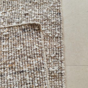 May include: A textured area rug featuring a chunky, looped weave in a neutral color palette of beige, cream, and dark brown. The rug is folded over to reveal the underside, showcasing a consistent, dense, and knotted construction throughout the surface.