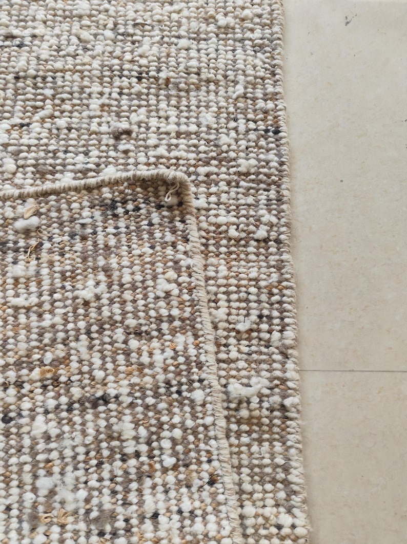May include: A textured area rug featuring a chunky, looped weave in a neutral color palette of beige, cream, and dark brown. The rug is folded over to reveal the underside, showcasing a consistent, dense, and knotted construction throughout the surface.