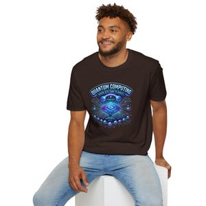 May include: A brown t-shirt featuring a vibrant graphic design related to quantum computing. The design includes the words "QUANTUM COMPUTING" in a curved banner, along with a circuit board-like illustration in shades of blue, purple, and teal. The text "WHERE BITS TURN TO QUBITS" is also included.