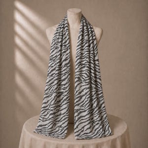 May include: A long rectangular scarf featuring a repeating black zebra stripe pattern printed on a light cream-coloured fabric. The scarf is draped vertically over a mannequin, displaying the full length of the fabric with its consistent animal print.