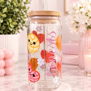 May include: A clear glass tumbler with a bamboo lid and a glass straw. The exterior features a colorful pattern of smiling concha bread icons, red and orange hearts, and small white flowers. The name Genesis is printed vertically in pink script.