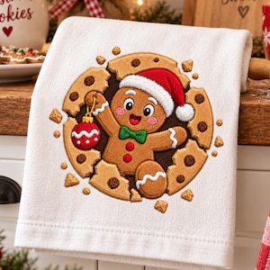 Puede incluir: Un paño de cocina blanco con un diseño bordado de una galleta de jengibre sonriente que lleva un gorro de Papá Noel rojo y blanco y una pajarita verde. La galleta de jengibre emerge de una galleta grande sosteniendo un adorno navideño rojo.