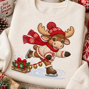 Puede incluir: Una sudadera de color crema con el gráfico de un alce marrón que lleva un suéter de punto rojo y blanco, un gorro con pompón a juego y una bufanda. El alce está patinando sobre hielo y tira de un trineo de madera con un regalo verde y cascabeles.