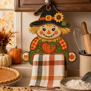 May include: A decorative fabric scarecrow towel holder featuring a black witch hat with a sunflower, a pumpkin graphic, and side accents of a candy corn and a maple leaf. A plaid kitchen towel in orange, green, and white hangs from the holder.