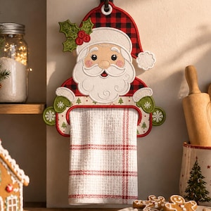 May include: A hanging kitchen towel holder shaped like Santa Claus, featuring a red and black buffalo plaid hat, a white beard, and green mittens. The holder displays a white waffle-weave towel with a red plaid pattern hanging from the centre.