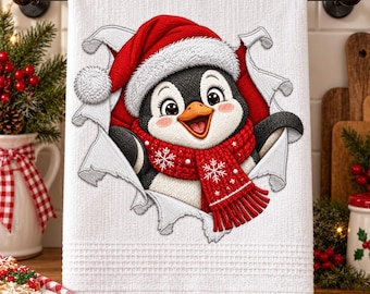 May include: A white waffle-weave kitchen towel featuring a central graphic of a cartoon penguin wearing a red Santa hat and a red knitted scarf with white snowflake patterns, appearing to burst through the fabric of the towel itself.