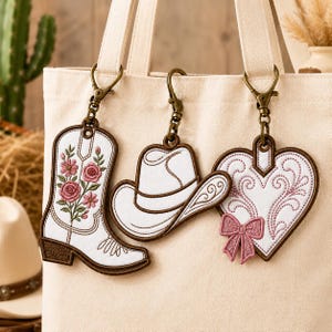 May include: Three embroidered bag charms attached to a cream canvas tote bag. The charms include a white cowboy boot with pink floral embroidery, a white cowboy hat with brown stitching, and a white heart with pink swirls and a pink bow.