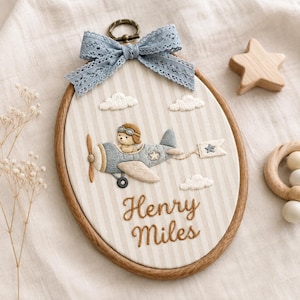 May include: An oval embroidery hoop featuring a beige and white striped fabric background. The design includes a stitched light blue aeroplane with a bear pilot wearing goggles, white clouds, and the embroidered brown text Henry Miles.