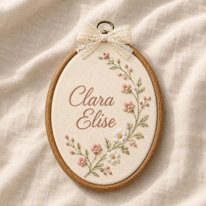 May include: An oval wooden embroidery hoop containing cream-colored fabric with the embroidered name Clara Elise in pink script. A floral vine with pink and white flowers and green leaves curves around the text, topped with a cream lace bow.