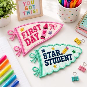 May include: Two embroidered felt pennants with school themes. One pink pennant reads FIRST DAY with a crayon graphic. One light green checkered pennant reads STAR STUDENT with a pencil graphic. Both feature decorative loops and embroidered stars.