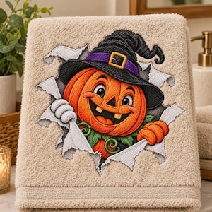 May include: A beige hand towel featuring an embroidered design of a smiling orange jack-o'-lantern wearing a black witch hat with a purple band and gold buckle. The pumpkin appears to be emerging from a tear in the fabric of the towel.