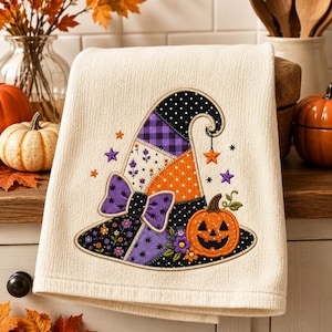 Può includere: Un canovaccio color crema con un ricamo raffigurante un cappello da strega in stile patchwork. Il cappello presenta motivi in tessuto viola, arancione e nero con pois, quadri e disegni floreali, arricchito da un fiocco viola e una zucca.