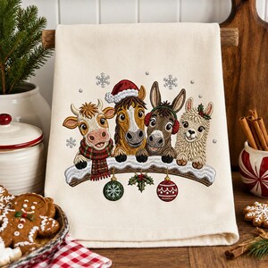 May include: A cream-colored kitchen towel featuring an embroidered design of a cow, horse, donkey, and llama resting on a snowy branch. The animals wear festive accessories like a Santa hat, earmuffs, and a plaid scarf, with hanging ornaments.