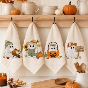 Può includere: Quattro strofinacci color bianco sporco appesi a una rastrelliera in legno, ognuno con un fantasma ricamato. I disegni includono un fantasma con una carriola, un girasole, dentro una zucca e uno che regge un cartello con scritto pumpkin patch.