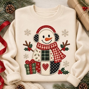 Puede incluir: Una sudadera de cuello redondo color crema con un gráfico central bordado de un muñeco de nieve. El muñeco lleva orejeras rojas, una bufanda de cuadros rojos y blancos, y un cuerpo de patchwork con motivos de acebo, lunares y corazones.