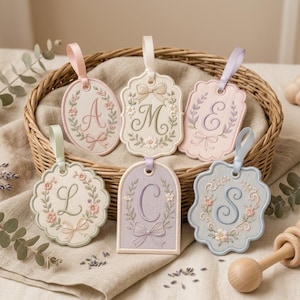 May include: Six fabric hanging tags with embroidered monograms A, M, E, L, C, and S. Each tag features floral wreaths, bows, and satin ribbon loops in pastel shades of pink, cream, lavender, and blue, with scalloped or arched edges.