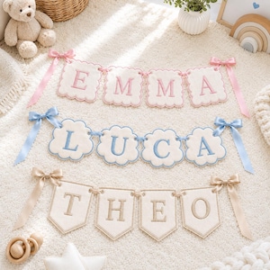 May include: Three personalized fabric banners featuring the names EMMA, LUCA, and THEO. The top banner has pink scalloped edges and pink lettering. The middle banner has blue cloud-shaped edges and blue lettering. The bottom banner has beige pennant-shaped edges and beige lettering, each with matching satin ribbon bows.