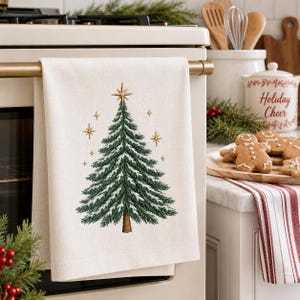 Pode incluir: Um pano de cozinha de cor creme com uma árvore de Natal bordada em verde, com ramos brancos cobertos de neve, um tronco castanho e detalhes de estrelas douradas. O pano está pendurado numa barra horizontal, exibindo um design festivo.