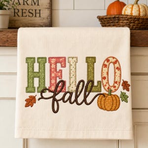 May include: A cream-colored kitchen towel featuring embroidered text that reads HELLO fall. The letters are filled with various patterns in shades of green, red, and cream. The design includes a small orange pumpkin and several autumn leaves.