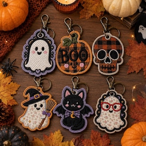 May include: Six embroidered keychains featuring Halloween themes: a white ghost with a purple border, an orange plaid pumpkin with the text BOO, a black and orange plaid skull, a ghost wearing a witch hat, a black cat with wings, and a ghost with red glasses.