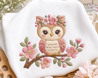 May include: A white fabric garment featuring an embroidered design of a brown owl with large eyes, wearing a pink floral crown. The owl is perched on a brown branch adorned with pink cherry blossoms and green leaves, centered on the white fabric.