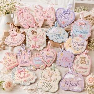 Puede incluir: Una colección de adornos de tela bordada en varias formas, incluyendo corazones, coronas, castillos y nubes. Cada pieza presenta colores pastel, colgadores de cinta de satén y nombres personalizados como Sophia, Amelia, Rosie, Charlotte, Ella, Lily, Theo, Clara, Bella y Emma.