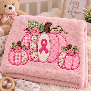 Könnte beinhalten: Eine weiche, rosafarbene Plüschdecke mit einer Stickerei von drei Kürbissen. Der mittlere Kürbis zeigt eine rosa Schleife zur Unterstützung der Brustkrebsvorsorge. Die Kürbisse sind mit Blumen-, Vichy- und Punktmustern in Rosatönen verziert.