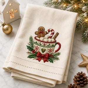 Puede incluir: Una toalla de tela color crema con un diseño bordado de una taza blanca llena de malvaviscos, un hombre de jengibre y un bastón de caramelo. La taza está decorada con patrones rojos y verdes, un lazo rojo y hojas de acebo verdes.