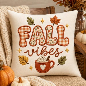 Peut inclure: Un coussin carré blanc cassé avec une broderie indiquant FALL VIBES. Le mot FALL alterne entre un motif vichy orange et un motif floral, tandis que vibes est en cursive. En dessous, une tasse avec un cœur et de la crème fouettée est visible.