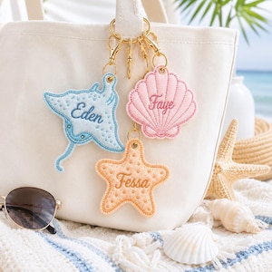 May include: Three embroidered felt keychains attached to a white canvas tote bag. A light blue stingray labeled Eden, a pink seashell labeled Faye, and an orange starfish labeled Fessa. Each keychain features gold-toned metal lobster clasps.