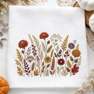 May include: A white fabric towel featuring a detailed embroidery design of various autumn-themed wildflowers, grasses, and leaves in shades of burnt orange, mustard yellow, deep red, and purple, arranged in a dense, horizontal botanical cluster.