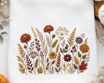 May include: A white fabric towel featuring a detailed embroidery design of various autumn-themed wildflowers, grasses, and leaves in shades of burnt orange, mustard yellow, deep red, and purple, arranged in a dense, horizontal botanical cluster.