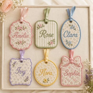 May include: Six fabric hanging tags with embroidered names and floral motifs. The tags feature various shapes including scalloped, rectangular, oval, and house-like silhouettes, each with a matching ribbon loop for hanging and delicate floral embroidery.