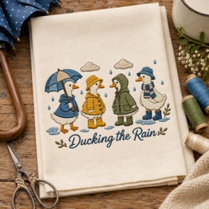 May include: A cream-coloured kitchen towel featuring an embroidered design of four white ducks wearing rain gear, including hats, coats, and boots, with the text "Ducking the Rain" in blue script, surrounded by embroidered raindrops and clouds.