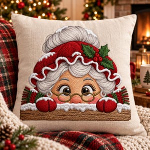 May include: A square beige throw pillow featuring an embroidered illustration of a character with white hair, round glasses, and a red hat with white trim and holly leaves. The character wears red mittens and rests hands on a brown wooden ledge.