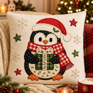Op de afbeelding: Een vierkant gebroken wit sierkussen met een geborduurde pinguïn die een rood-witte muts met stippen en een rood-wit geblokte sjaal draagt. De pinguïn houdt een groen geruit cadeau vast, omringd door geborduurde sneeuwvlokken en sterren.
