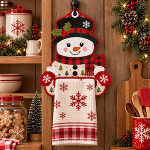 May include: A hanging kitchen towel holder shaped like a snowman wearing a black top hat with holly, a red and black buffalo plaid scarf, and red mittens. The attached white waffle-weave towel features red snowflake patterns and a plaid border.