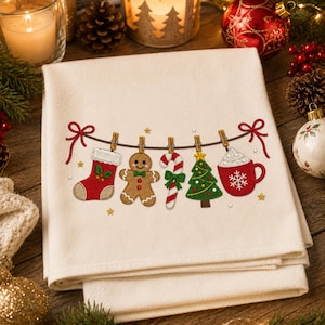 May include: A cream-colored kitchen towel featuring an embroidered design of a clothesline with red bows at each end. The line holds a red stocking, a gingerbread man, a candy cane, a green Christmas tree, and a red mug with a snowflake.